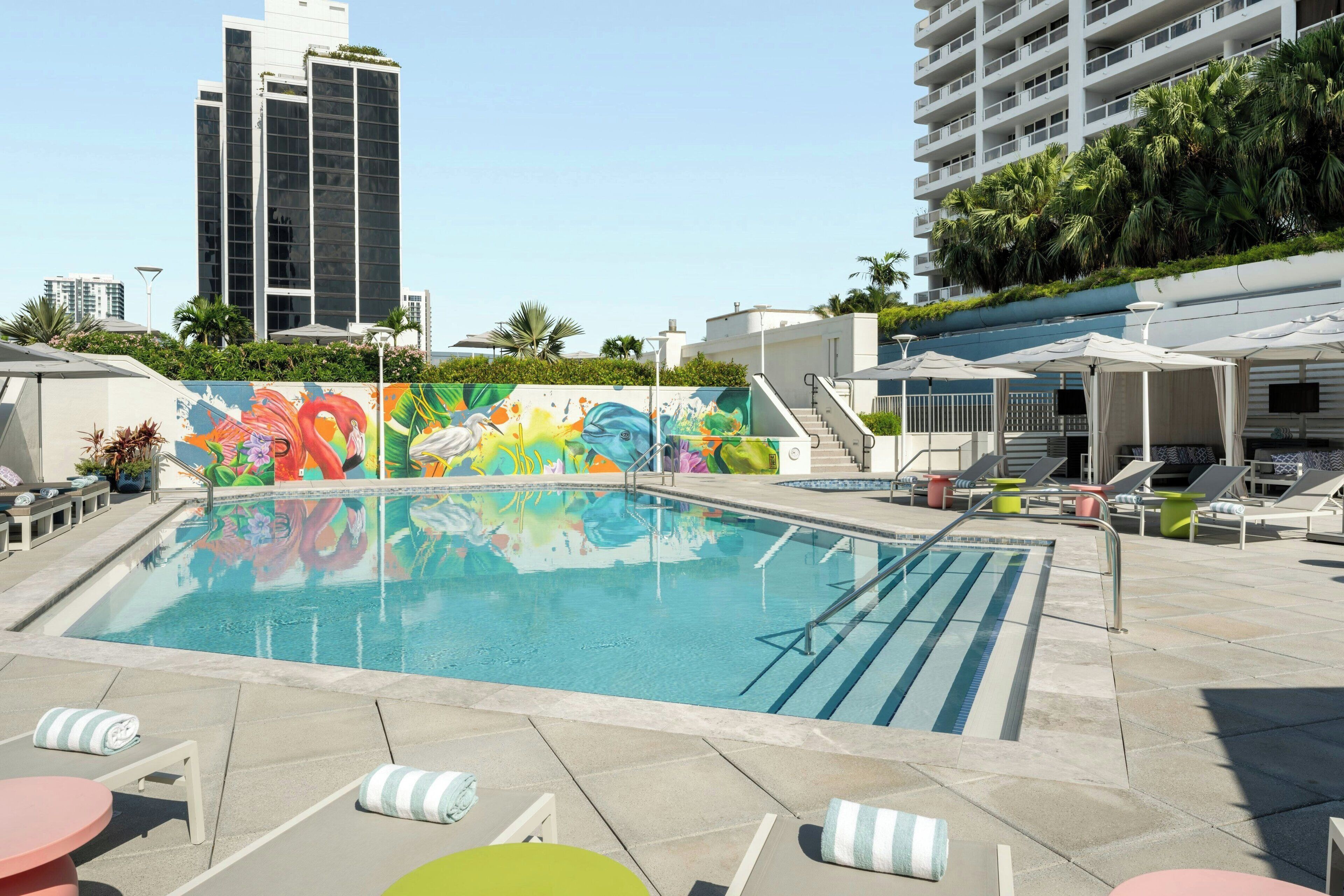 A rooftop pool with a colorful mural, lounge chairs, and a view of tall buildings.