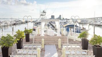 A wedding ceremony setup with white chairs and a table, overlooking a marina with boats and a city skyline.