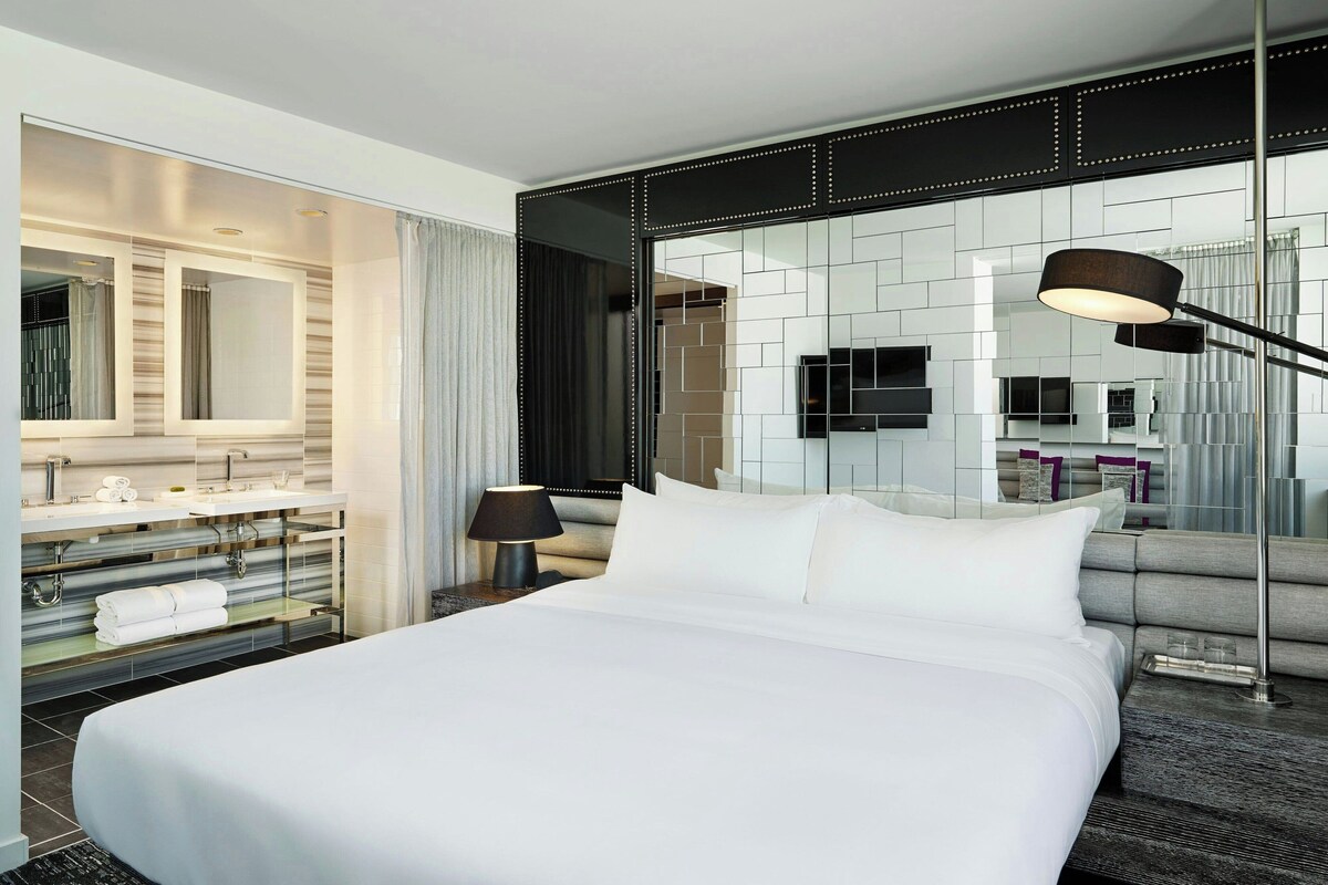 Ein modernes Hotelzimmer mit einem großen Bett, einem Schminktisch mit Spiegel, einem Flachbildfernseher und einem großen Fenster mit Vorhängen.