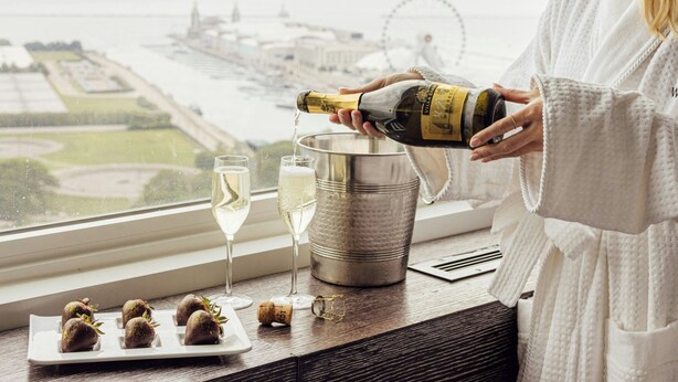 A person pouring champagne into a glass near a window overlooking a cityscape with a Ferris wheel. - The Wade (Chicago)