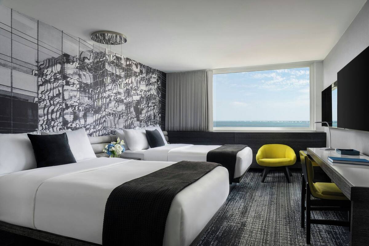Une chambre d’hôtel avec deux lits, un bureau, une chaise, une grande fenêtre donnant sur un beau paysage et un décor mural moderne.