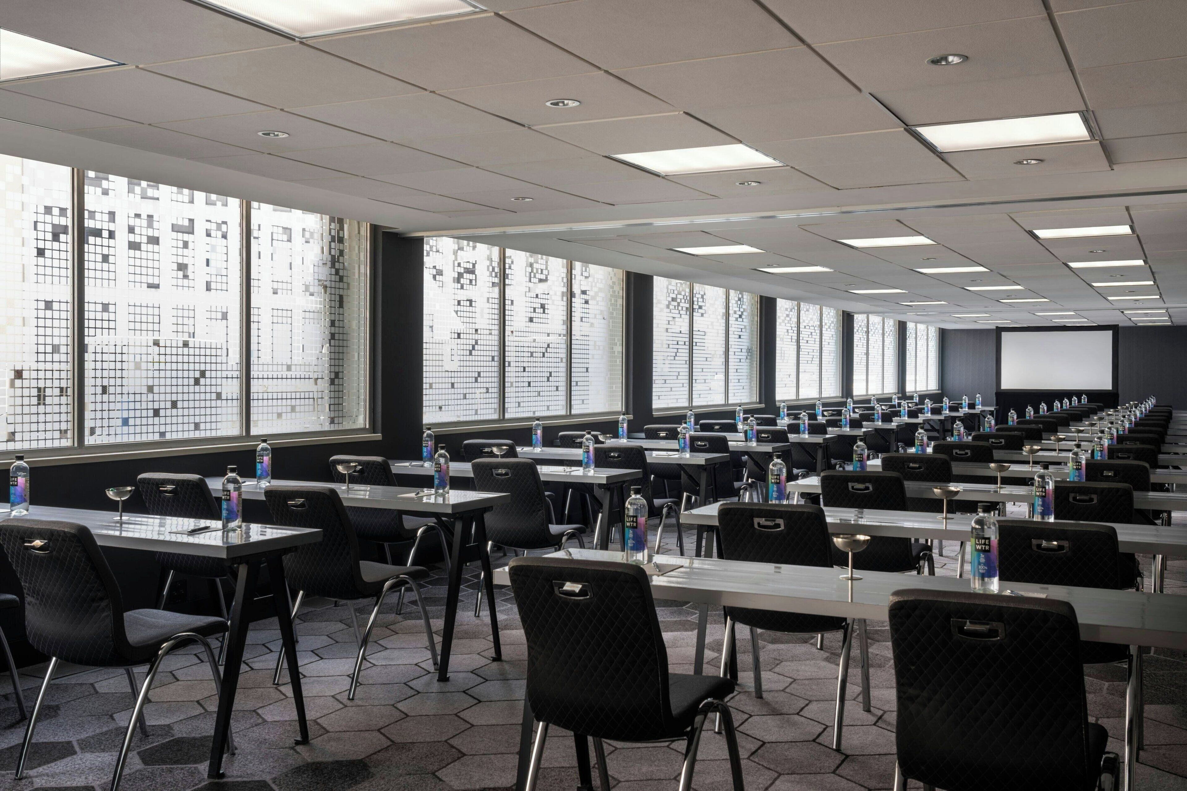 A conference room with multiple tables and chairs, large windows with a grid pattern, and a projector screen at the front.