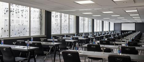 A conference room with multiple tables and chairs, large windows with a grid pattern, and a projector screen at the front.