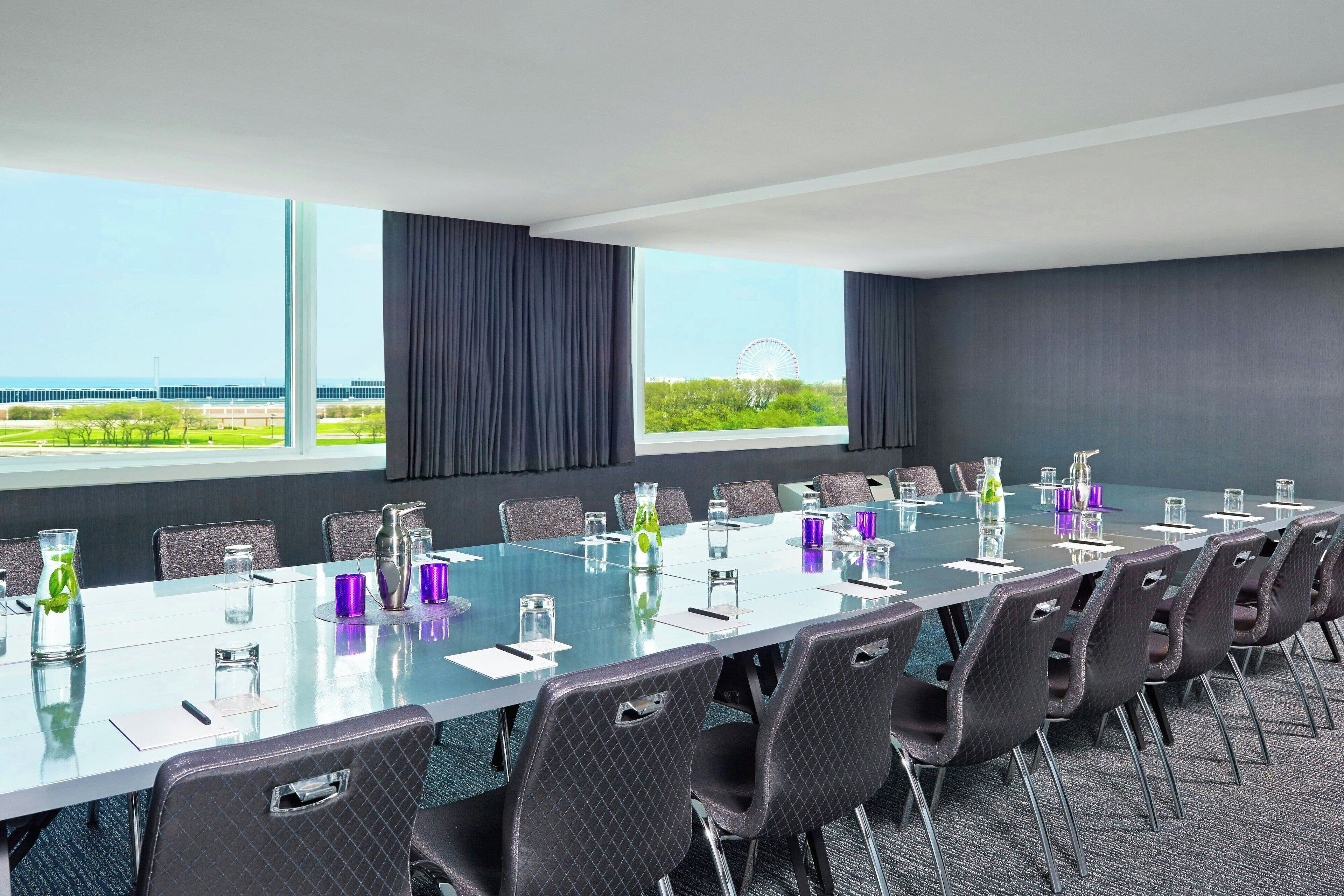 Un salón de conferencias con una larga mesa, sillas y grandes ventanales que ofrecen vistas a un edificio lejano y vegetación.