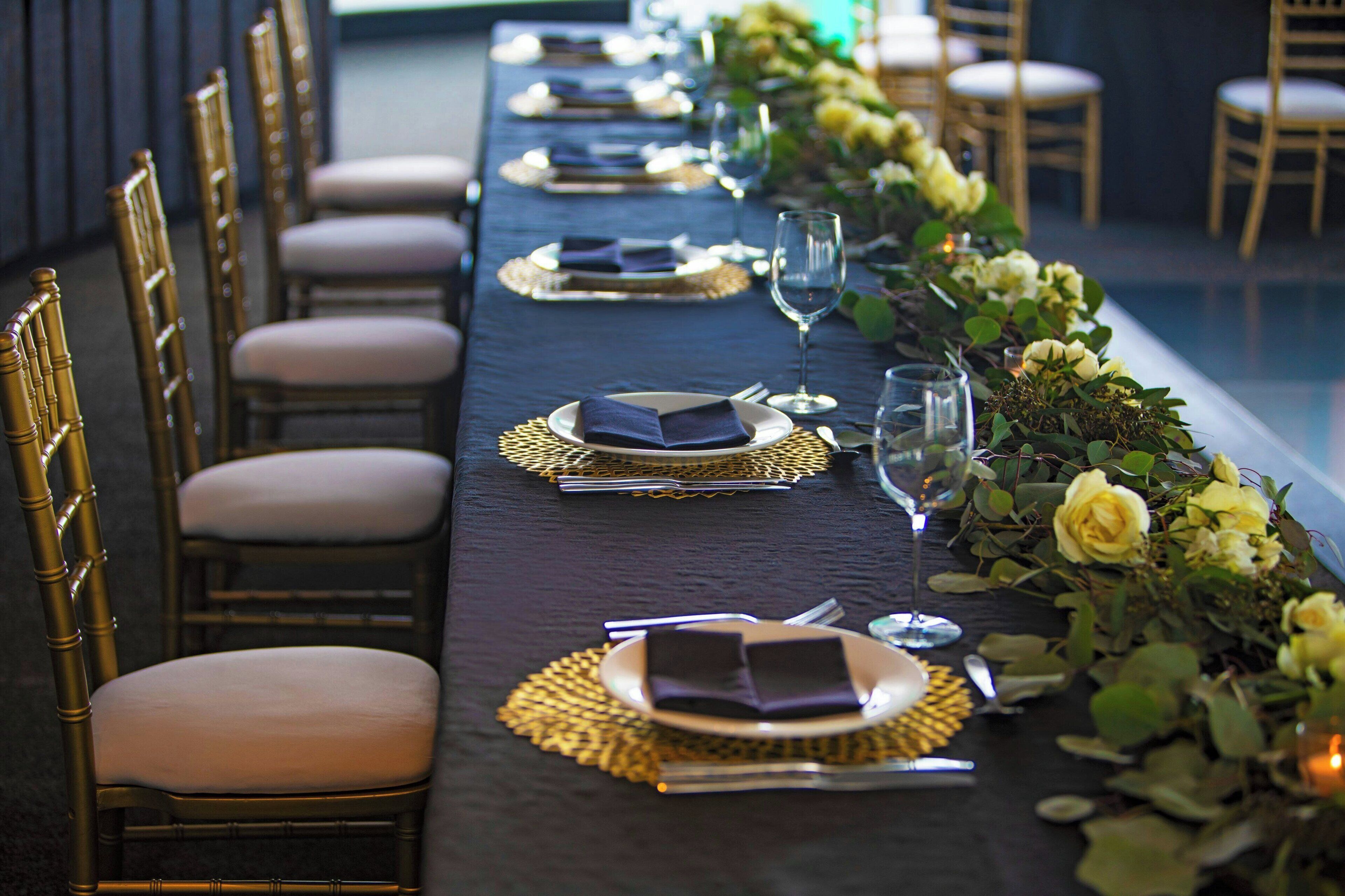 A long dining table with gold chairs, set with plates, napkins, and glasses, decorated with flowers and greenery.