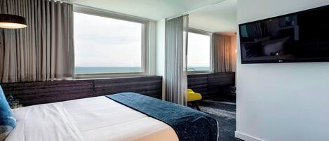 A hotel room with a bed, a TV, a desk, and a view of the sea.