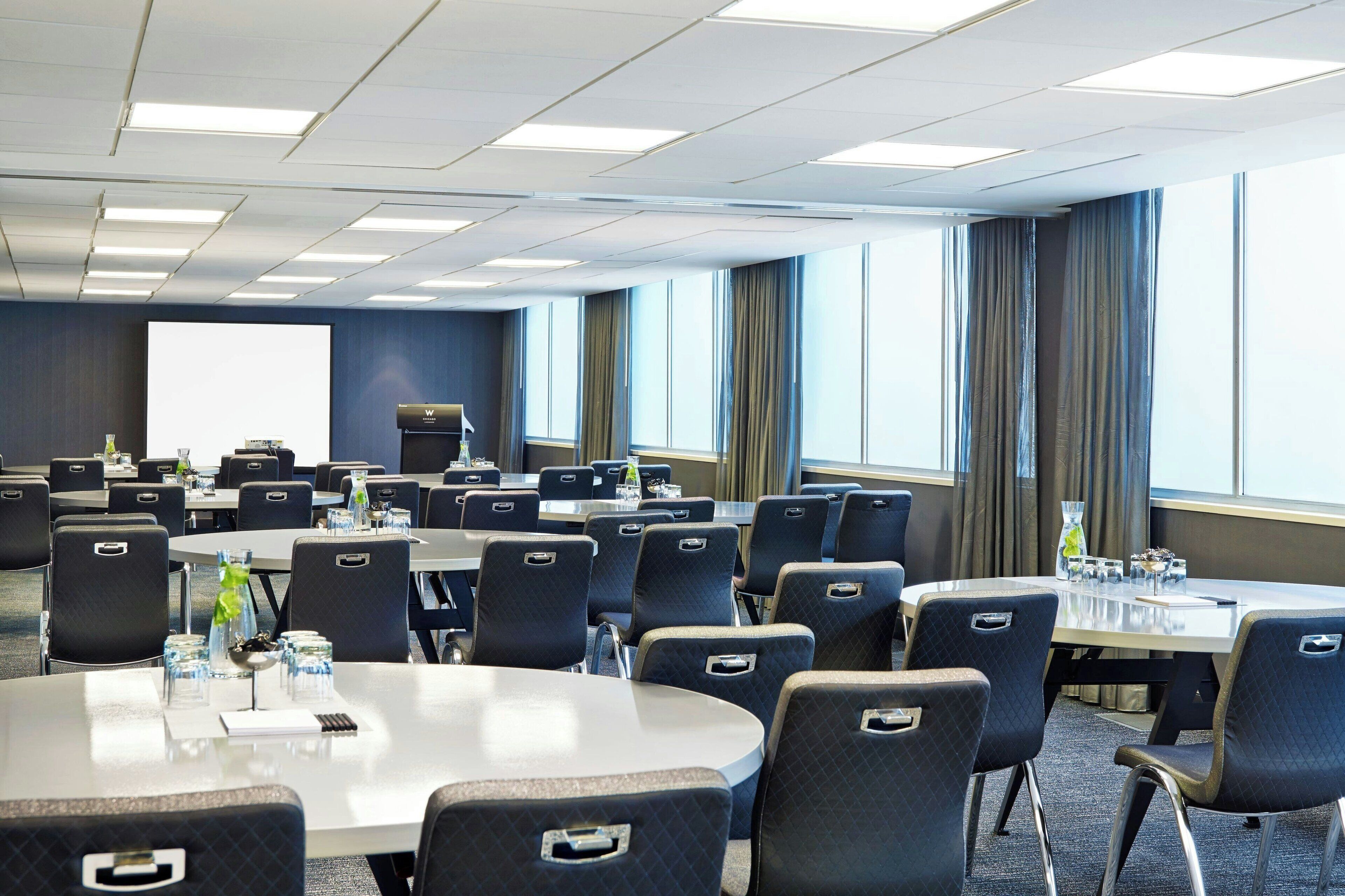 A conference room with multiple tables and chairs, large windows with curtains, and a projector screen.
