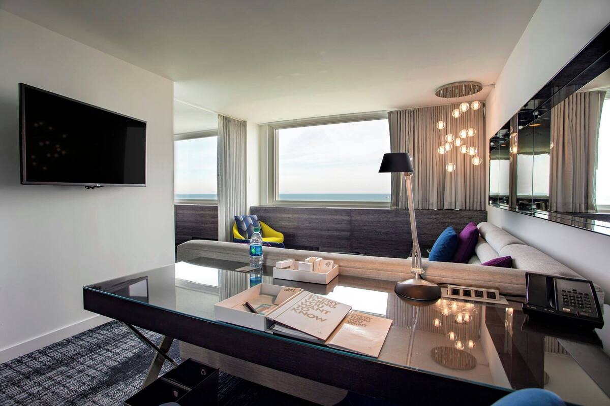 Une chambre d’hôtel moderne dotée d’un bureau en verre, d’un téléviseur à écran plat, d’un canapé avec des coussins, d’une grande fenêtre offrant une vue dégagée et d’un lustre.
