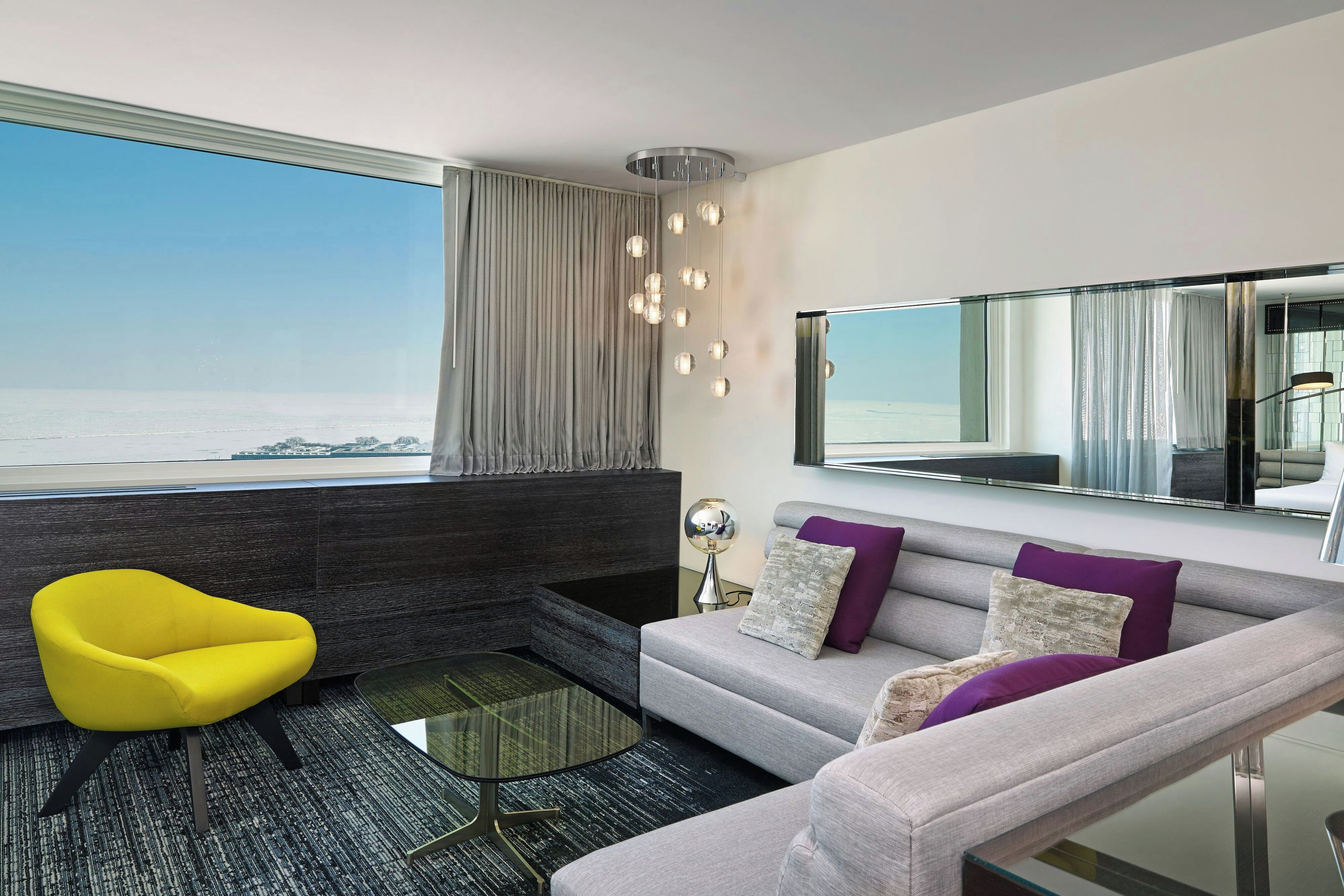 A modern living room with a sofa, a yellow chair, a glass coffee table, and a view of the sea.