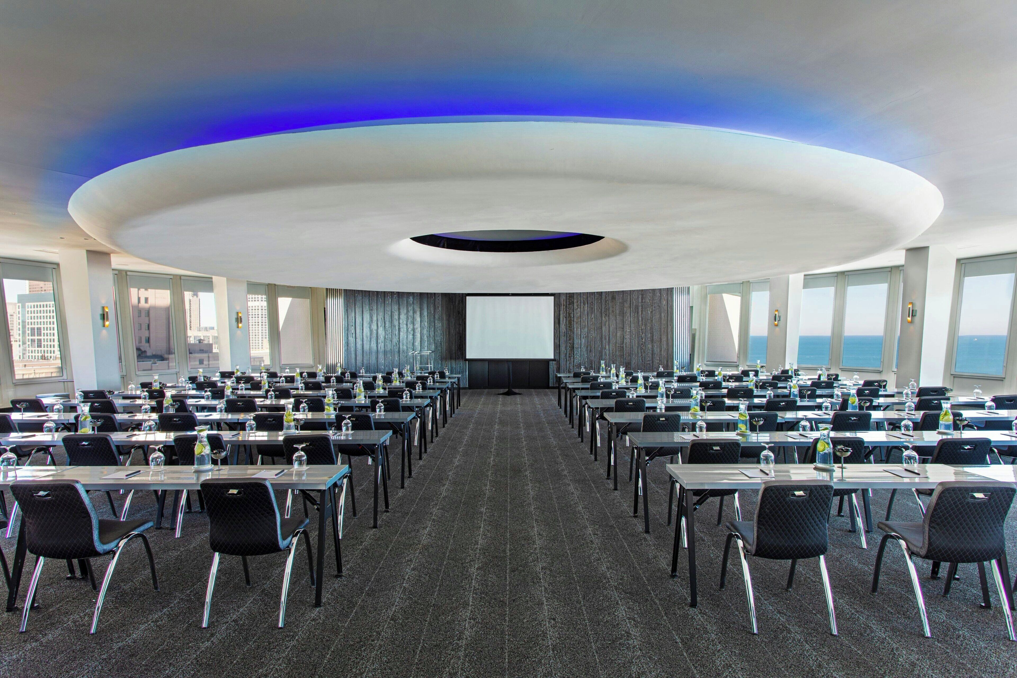 A conference room with a large screen at the front, multiple tables and chairs arranged for meetings, and large windows offering a view of the cityscape.