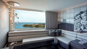 A modern hotel room with a large window offering a view of the sea and a cityscape. There is a seating area with a sofa and a small table. - The Wade (Chicago)