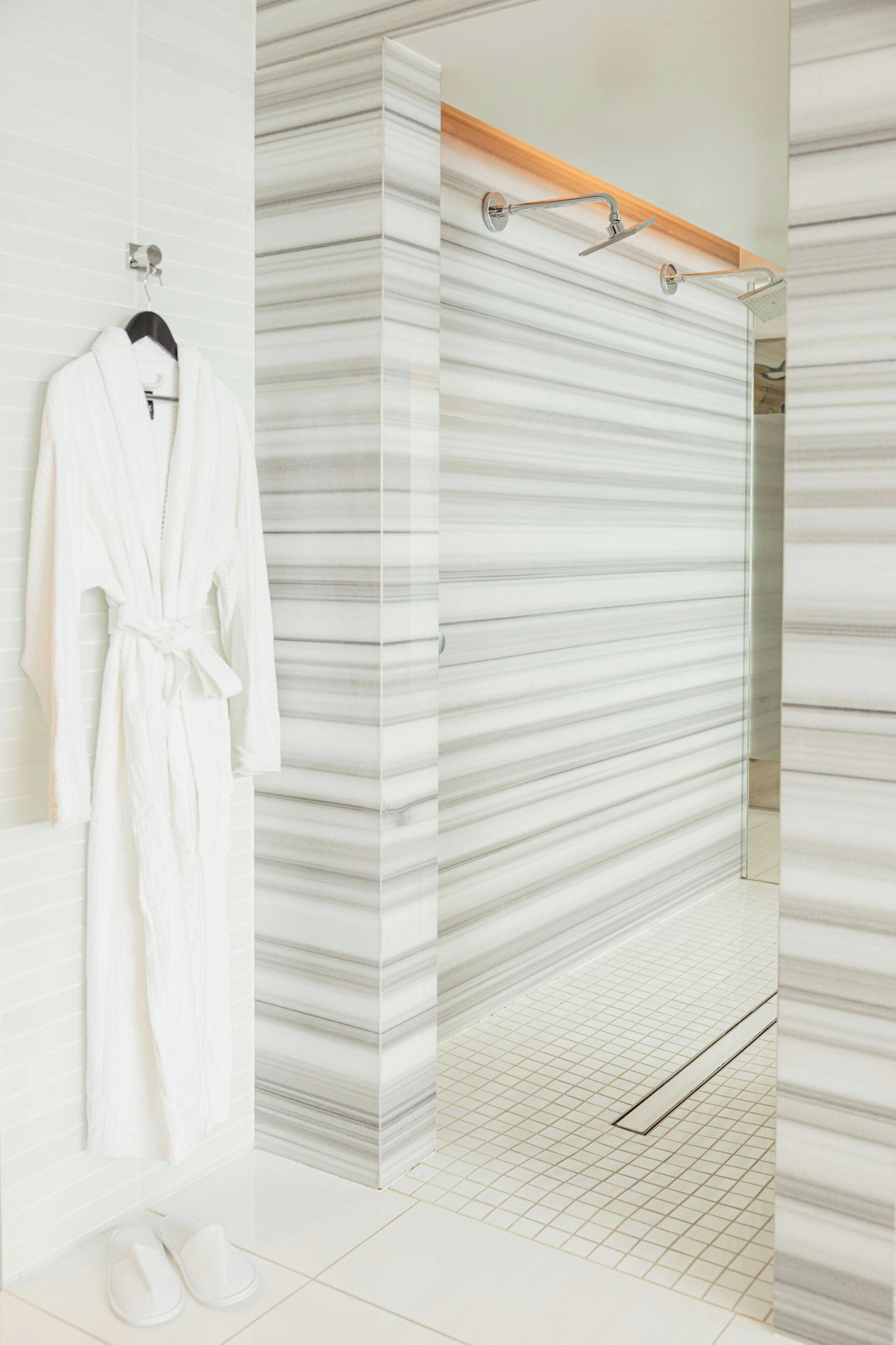 A bathroom with a marble shower wall, a hanging bathrobe, and a pair of slippers on the floor.