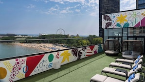 A rooftop terrace with a view of a beach and a Ferris wheel, featuring lounge chairs and a colorful mural. - The Wade (Chicago)