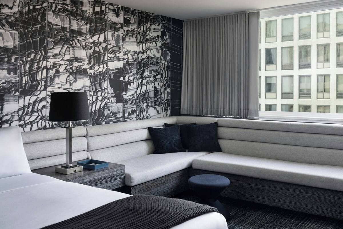 Une chambre d’hôtel moderne, dotée d’un canapé d’angle, d’une petite table ronde, d’une lampe et offrant une vue sur la ville grâce à de grandes fenêtres.