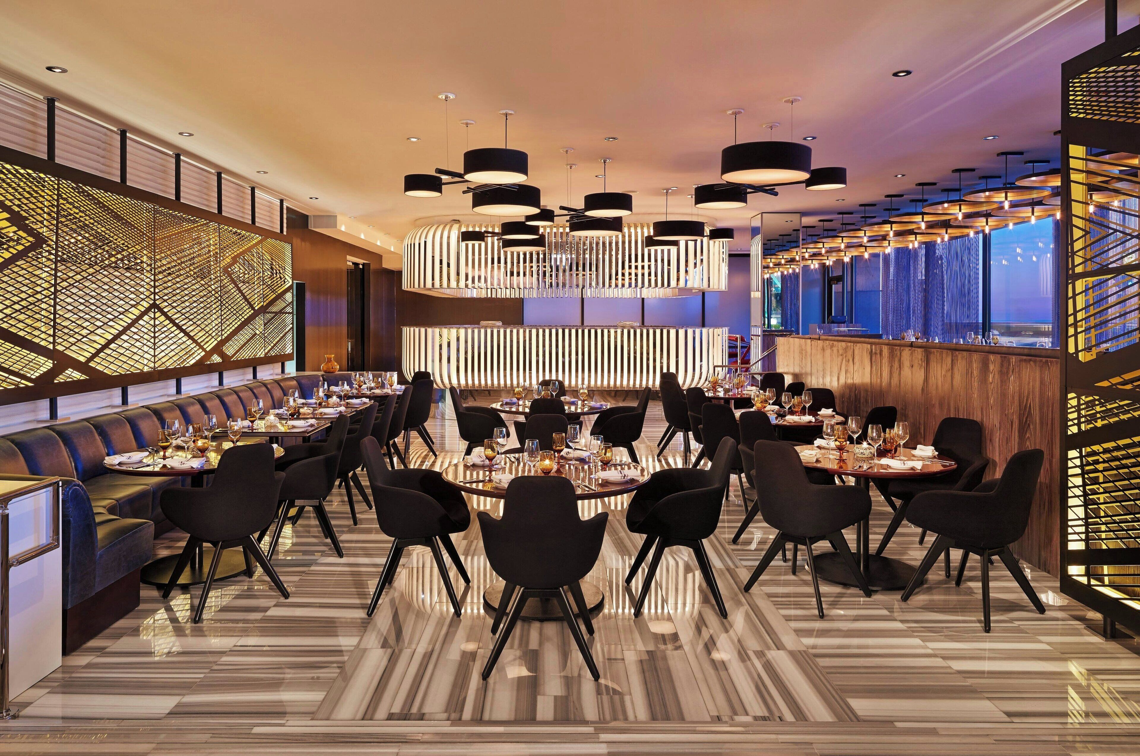 A modern dining area with multiple tables and chairs, a patterned floor, and contemporary lighting fixtures.