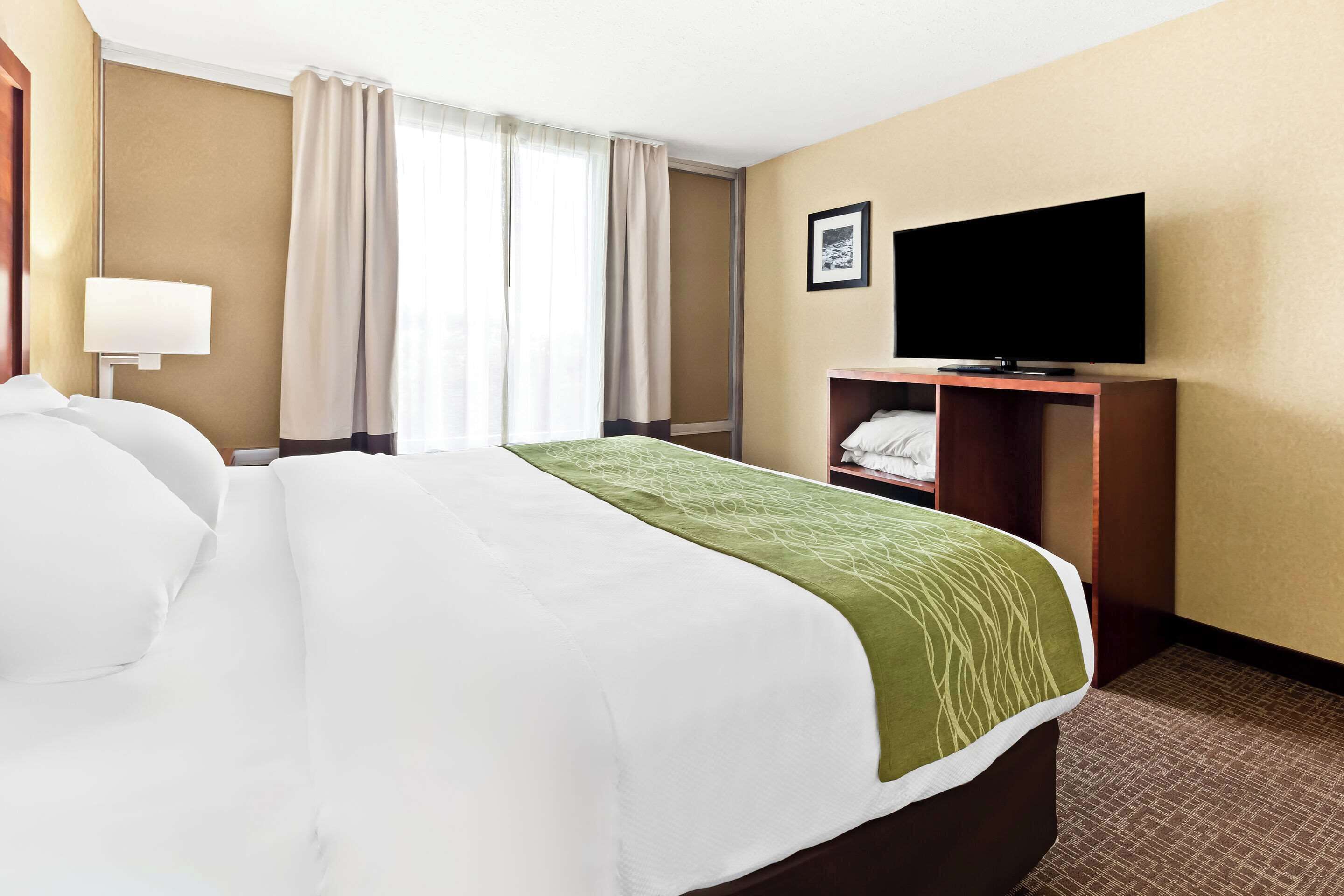 Una habitación de hotel con una cama grande, un televisor sobre un soporte de madera y una ventana con cortinas.