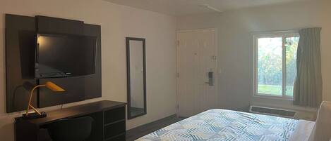 A hotel room with a bed, a desk, a chair, a TV, a mirror, and a window with curtains.