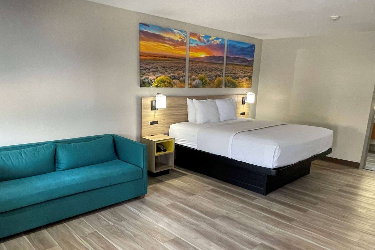 A hotel room with a bed, a teal sofa, a nightstand, and a wall-mounted painting of a sunset.
