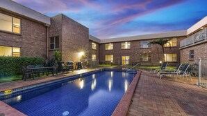 Piscina com borda de tijolos e espreguiçadeiras, cercada por um edifício com várias janelas e fachada de tijolos.