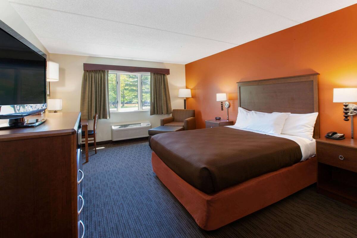Ein Hotelzimmer mit einem großen Bett, einem Schreibtisch mit Fernseher, einem Sessel, einer Schreibtischlampe und einem Fenster mit Vorhängen.
