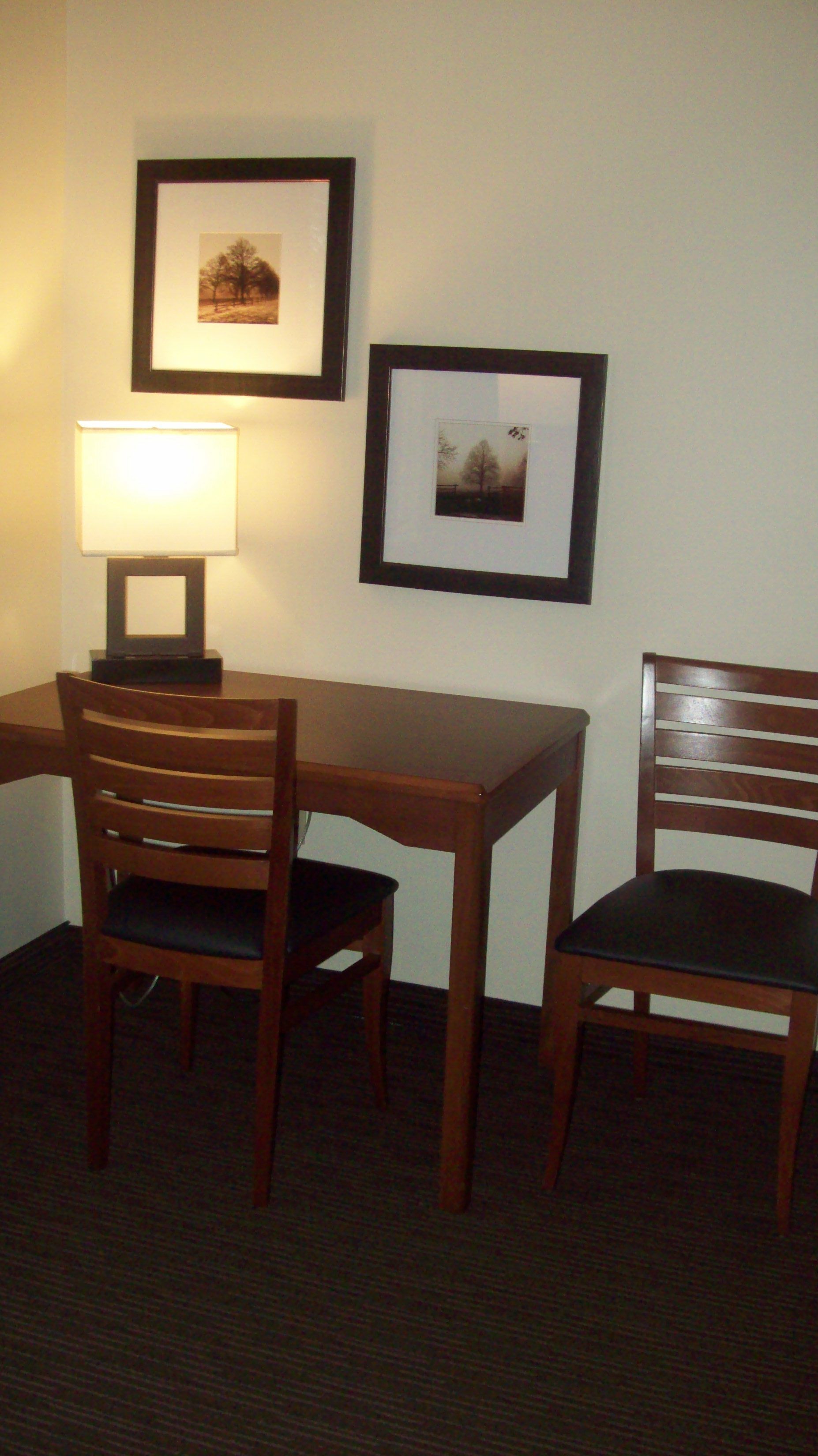 A small dining area with a table and two chairs, a lamp, and two framed pictures on the wall.