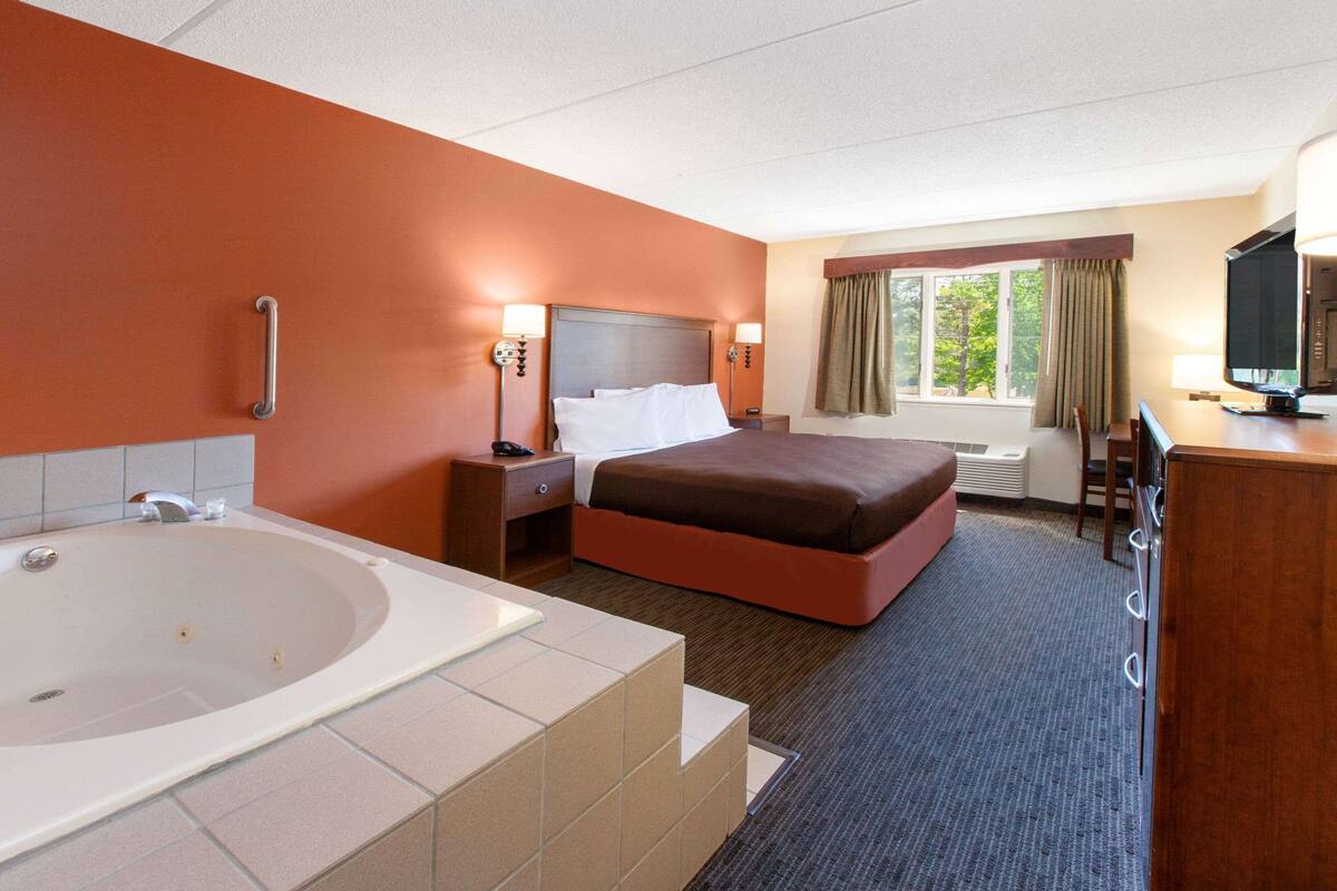 Ein Hotelzimmer mit einer großen Badewanne, einem Bett mit brauner Kopfteil, einem Schreibtisch mit Fernseher und einem Fenster mit Vorhängen.