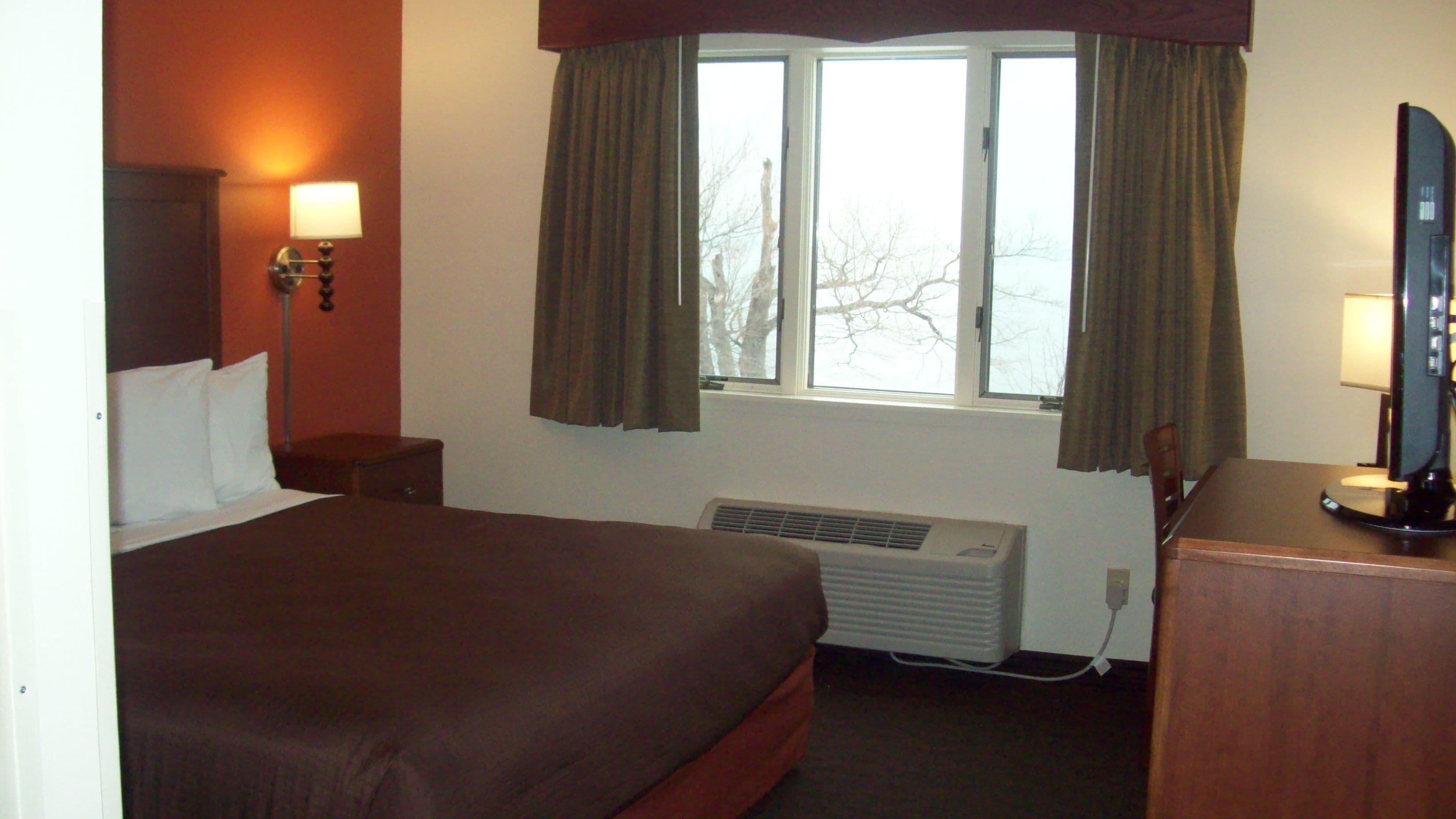 A hotel room with a large bed, a desk with a computer, a window with curtains, a wall-mounted lamp, and a heater.