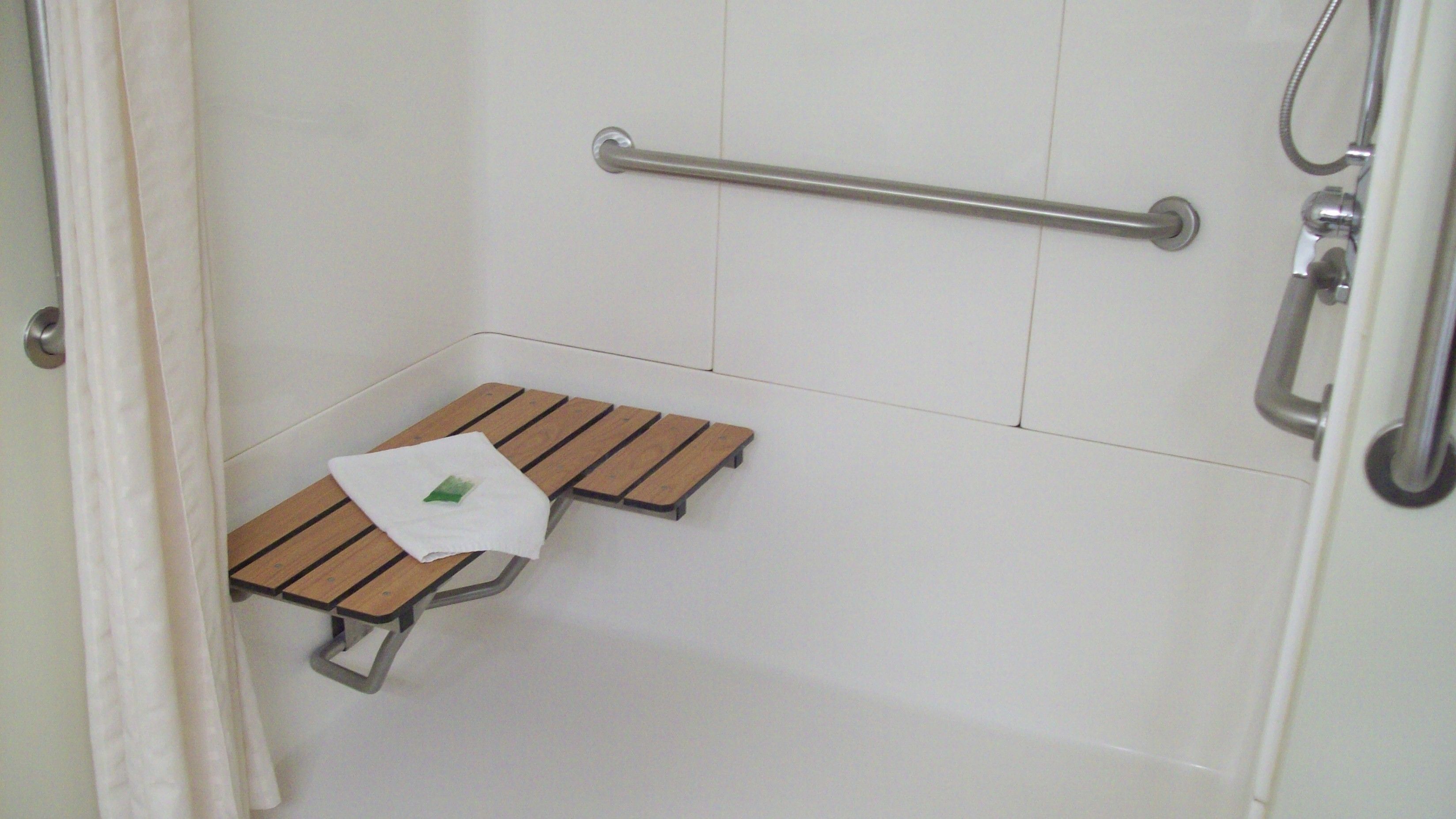 Ein Badezimmer mit weiß gefliestem Boden, einer Holzbank, einer Handstange und einem Duschbereich.