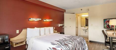 A hotel room with a bed, bedside table, lamp, desk, chair, and a bathroom visible through an open door.