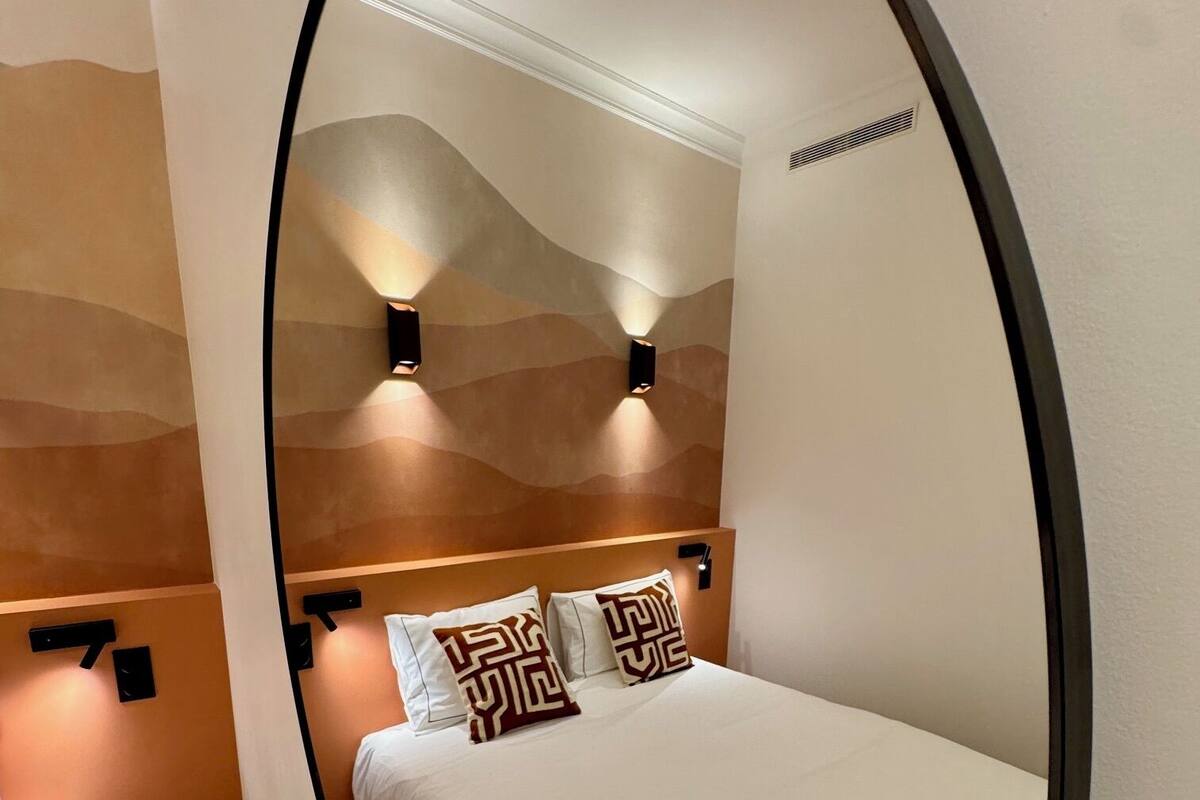 A bedroom with a bed, two wall-mounted lights, and a mirror reflecting the room.
