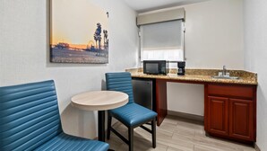 Quarto de hotel compacto com cozinha compacta, área de refeições com cadeiras estofadas em azul, uma pequena mesa redonda e um quadro de uma paisagem de pôr do sol na parede.