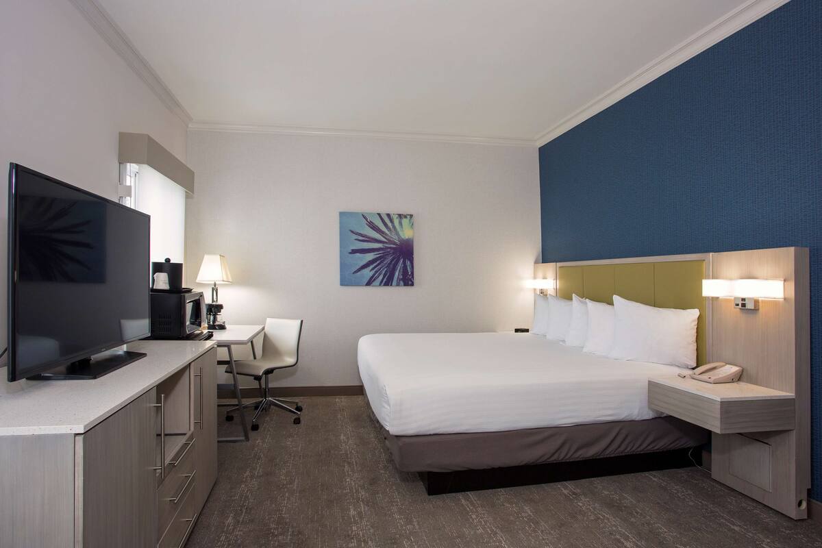 Ein Hotelzimmer mit Bett, Nachttisch, Lampe, Schreibtisch mit Computer, Fernseher und einem Bild an der Wand.