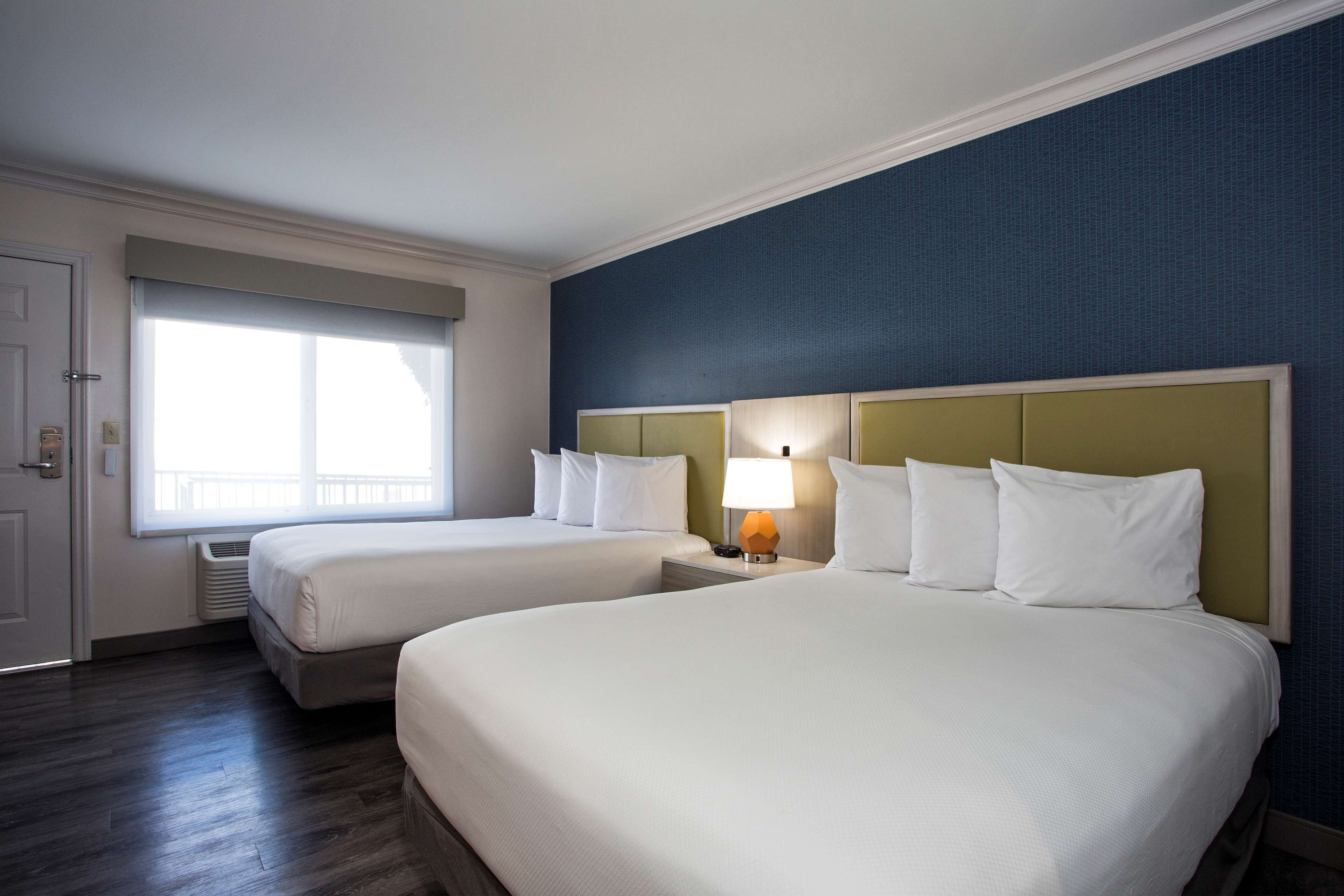 A hotel room with two beds, a window with blinds, a lamp on the nightstand, and a door.
