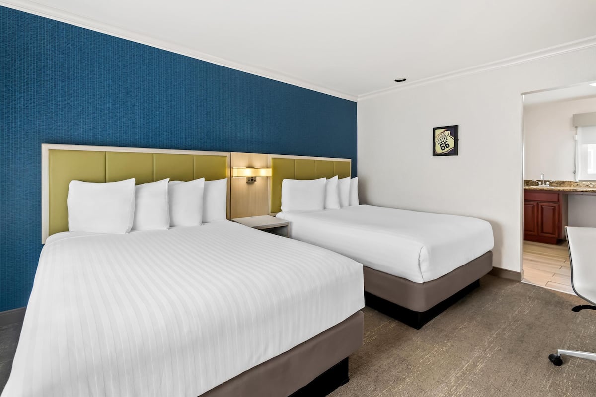 Ein Hotelzimmer mit zwei Betten, einem Nachttisch, einer Wanduhr und einer kleinen Küchenzeile.