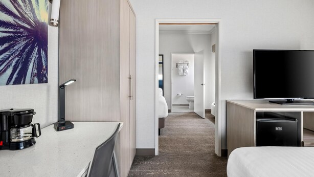 Quarto de hotel com cama, televisão, cafeteira e escrivaninha. Uma porta dá acesso a um banheiro e a um corredor.