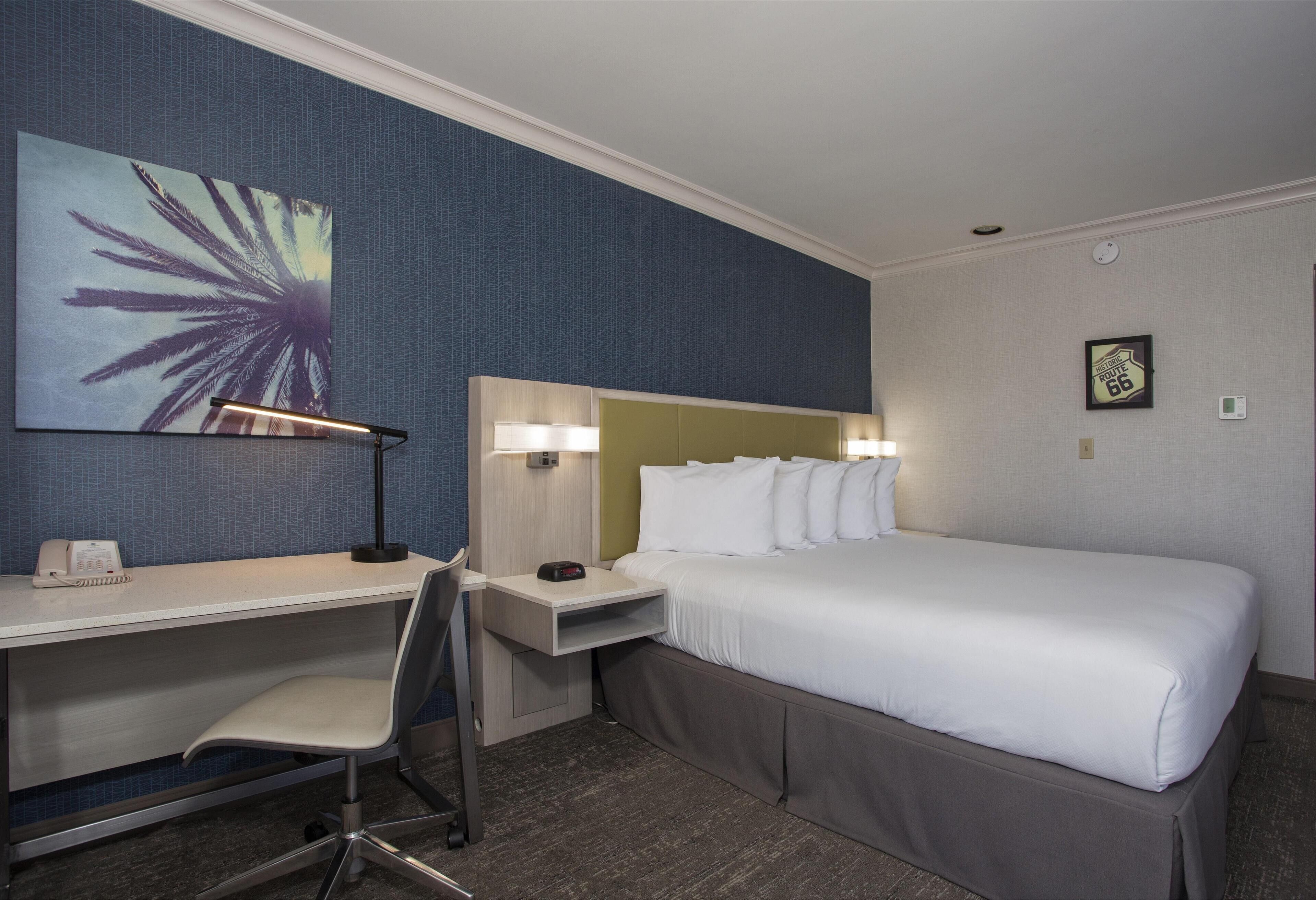 Quarto de hotel com cama, escrivaninha, cadeira e uma grande obra de arte na parede.