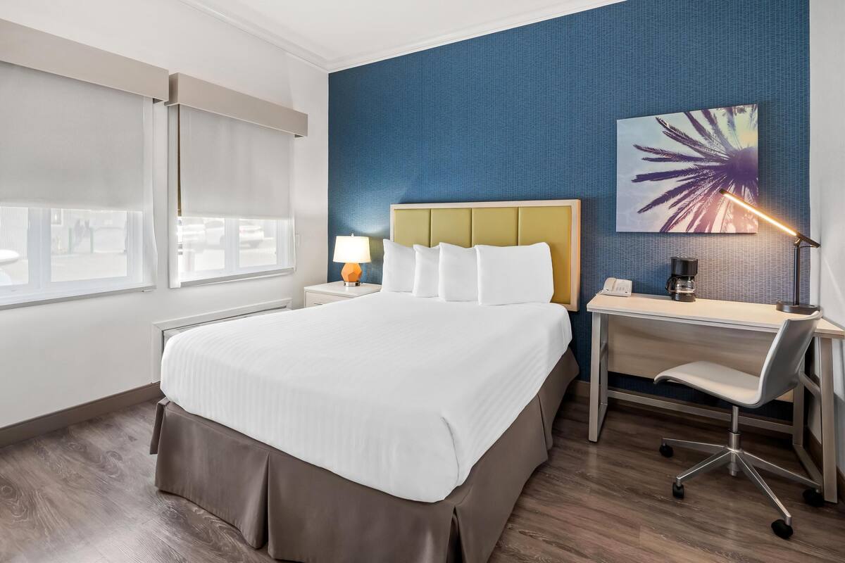 Ein Hotelzimmer mit einem großen Bett, einem Schreibtisch mit Stuhl, einer Lampe und einem Bild an der Wand.