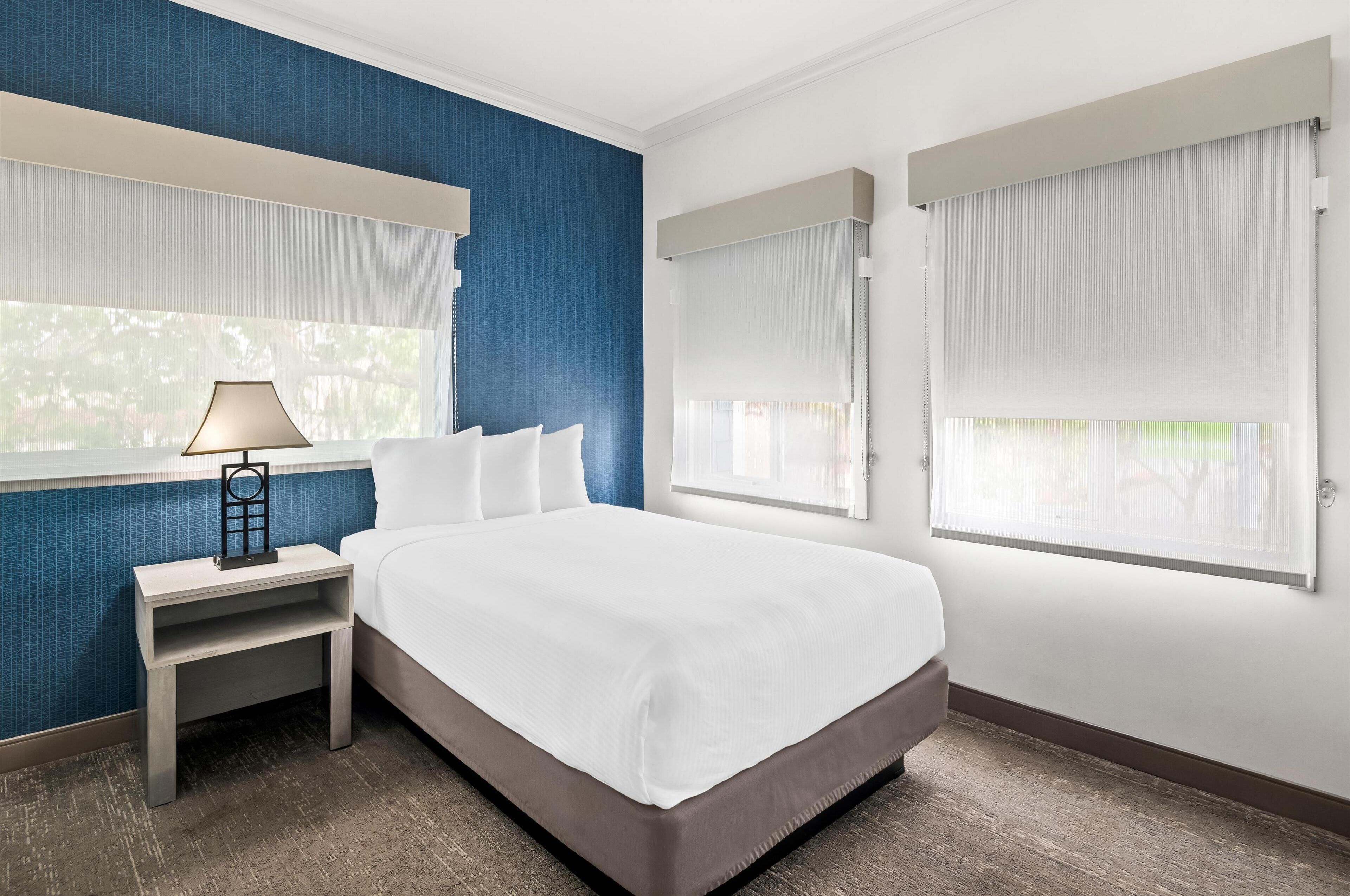 Une chambre d’hôtel comprenant un lit, une table de chevet, une lampe et deux fenêtres équipées de stores.