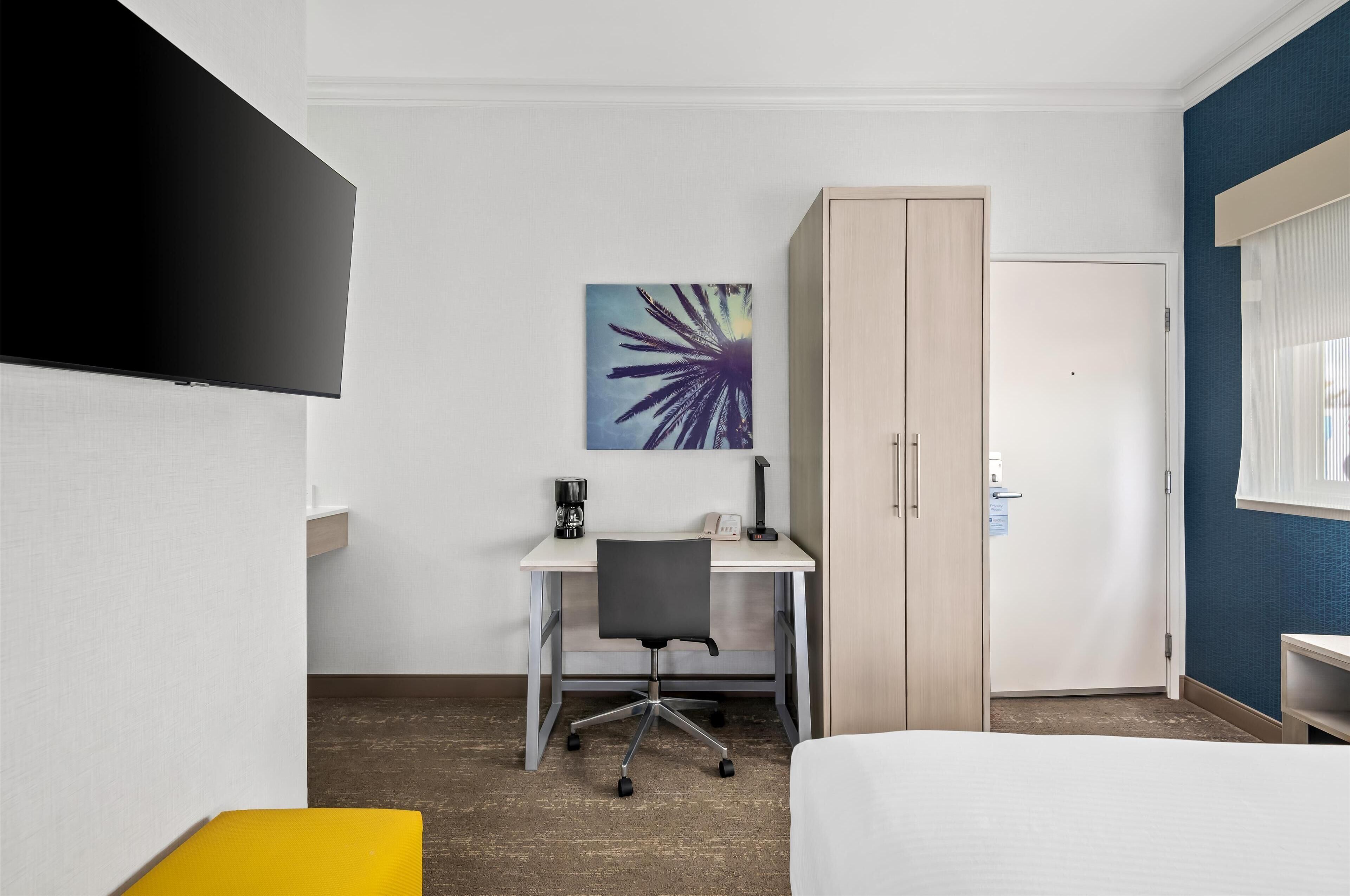 Une chambre d’hôtel comprenant un lit, un bureau avec une chaise, une armoire, un tableau au mur et un téléviseur fixé au mur.