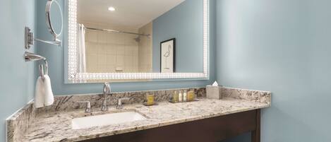 A bathroom with a marble sink, a large mirror with lights, a towel rack, and a framed picture on the wall.