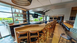 A modern bar area with a long counter, wooden chairs, and a view of greenery outside. - DoubleTree by Hilton Tampa Rocky Point Waterfront (Tampa)