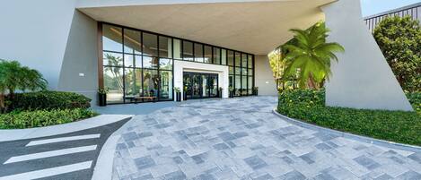 A modern building entrance with a curved pathway, large windows, and a palm tree.