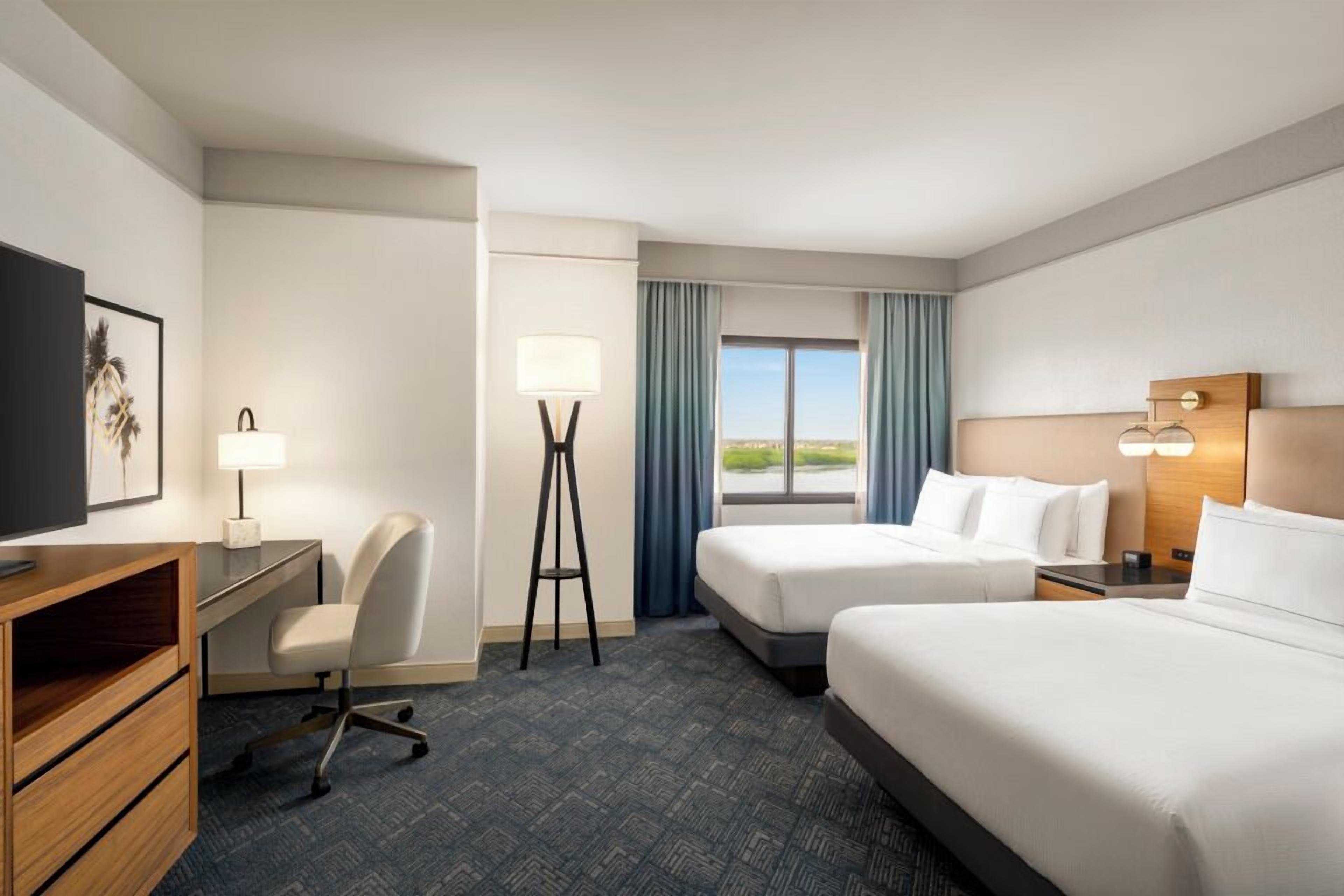 A hotel room with two beds, a desk, a chair, a TV, and a window with a view.