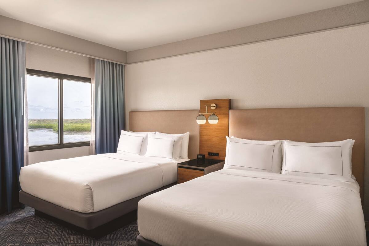A hotel room with two beds, a nightstand, a wall-mounted lamp, and a large window with curtains.