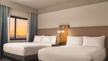 Quarto de hotel com duas camas, uma mesa de cabeceira, uma luminária fixada na parede e vista para o pôr do sol pela janela.