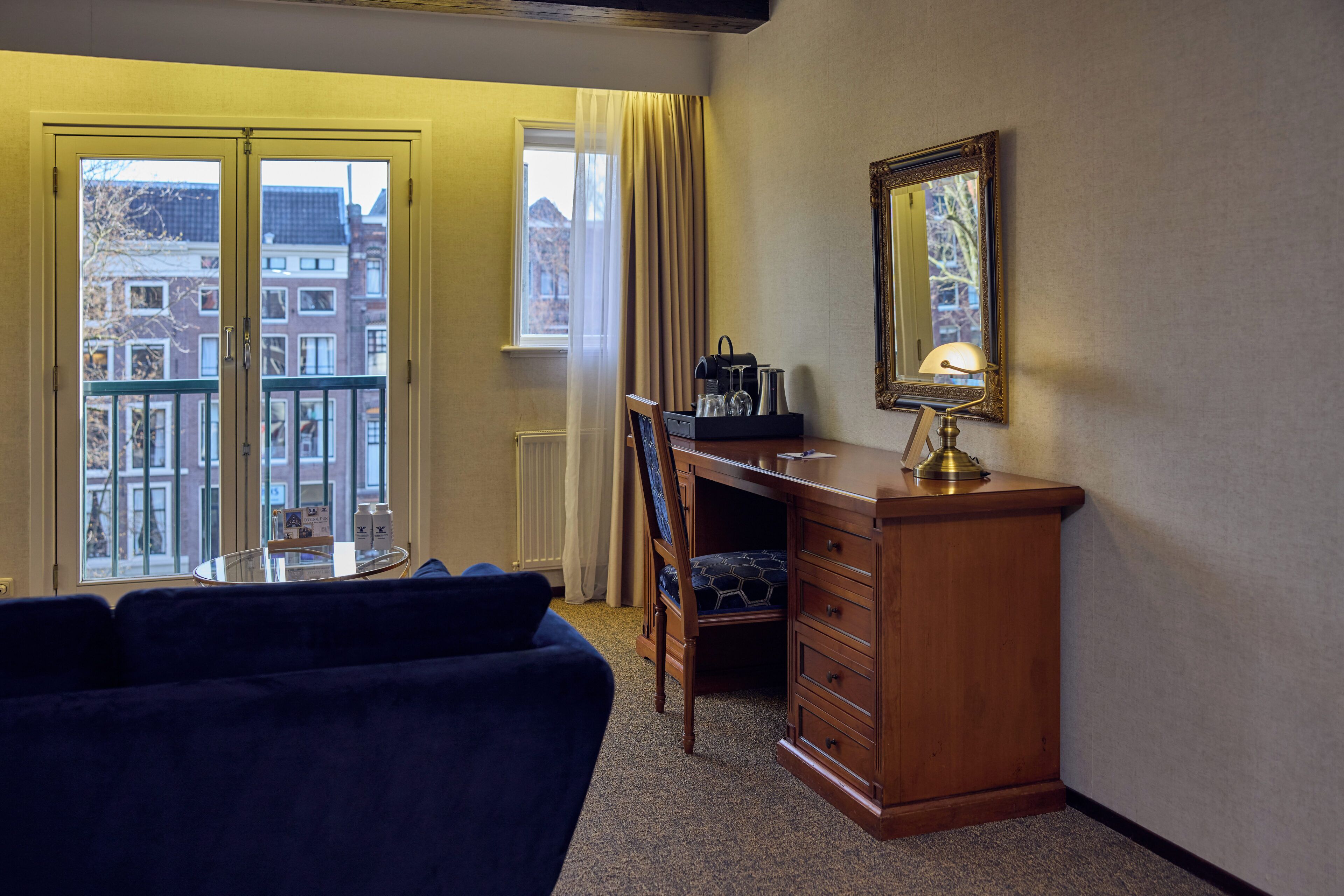 A hotel room with a balcony, a desk with drawers, a chair, a round table, and a mirror on the wall.
