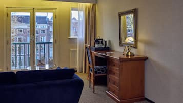 A hotel room with a balcony, a desk with drawers, a chair, a round table, and a mirror on the wall.