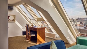 Une pièce avec un plafond en pente, des fenêtres de toit, un bureau, des chaises et une vue sur le paysage urbain.