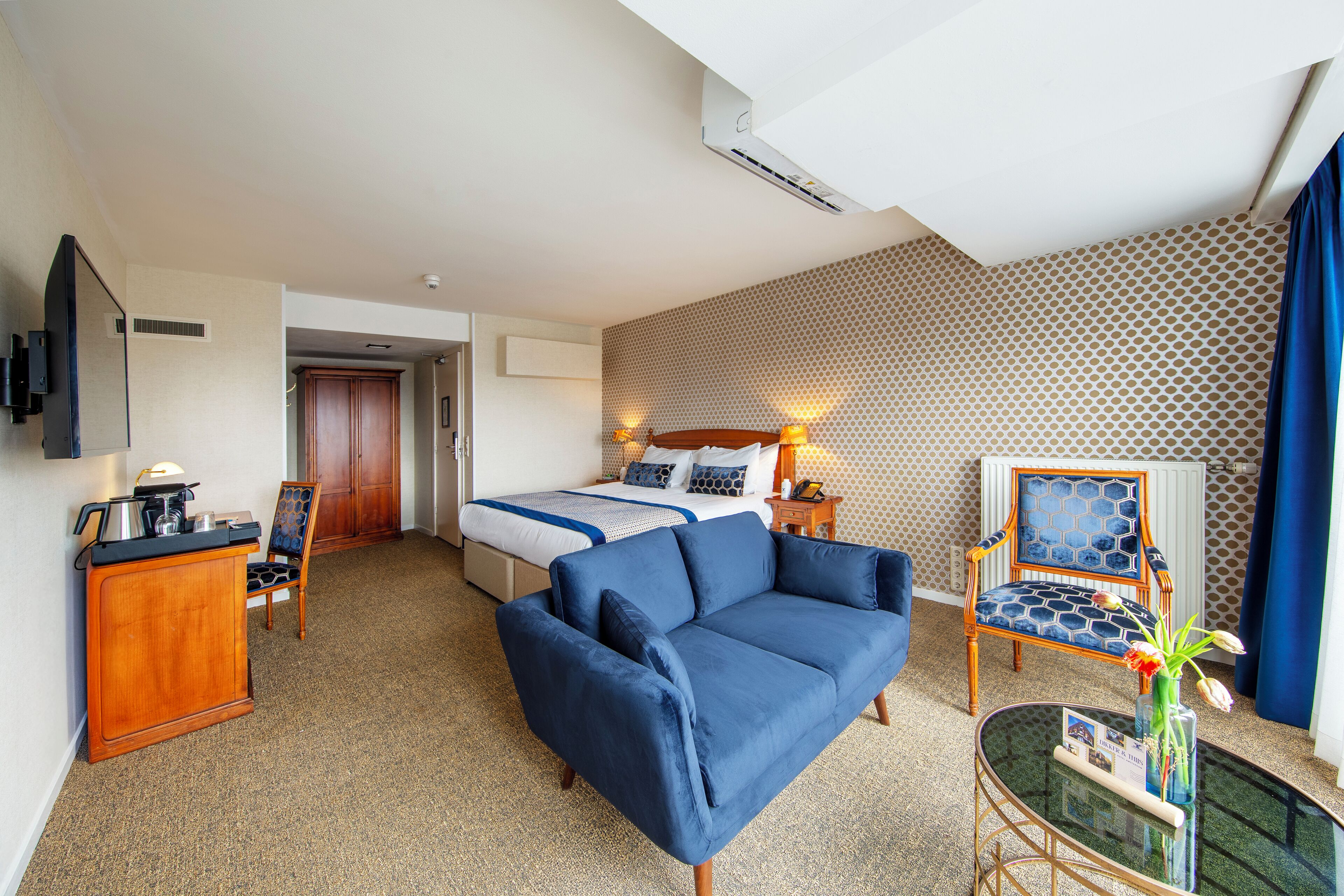 Habitación de hotel con cama, sofá azul, aparador de madera, una mesita con un jarrón y pared con diseño.