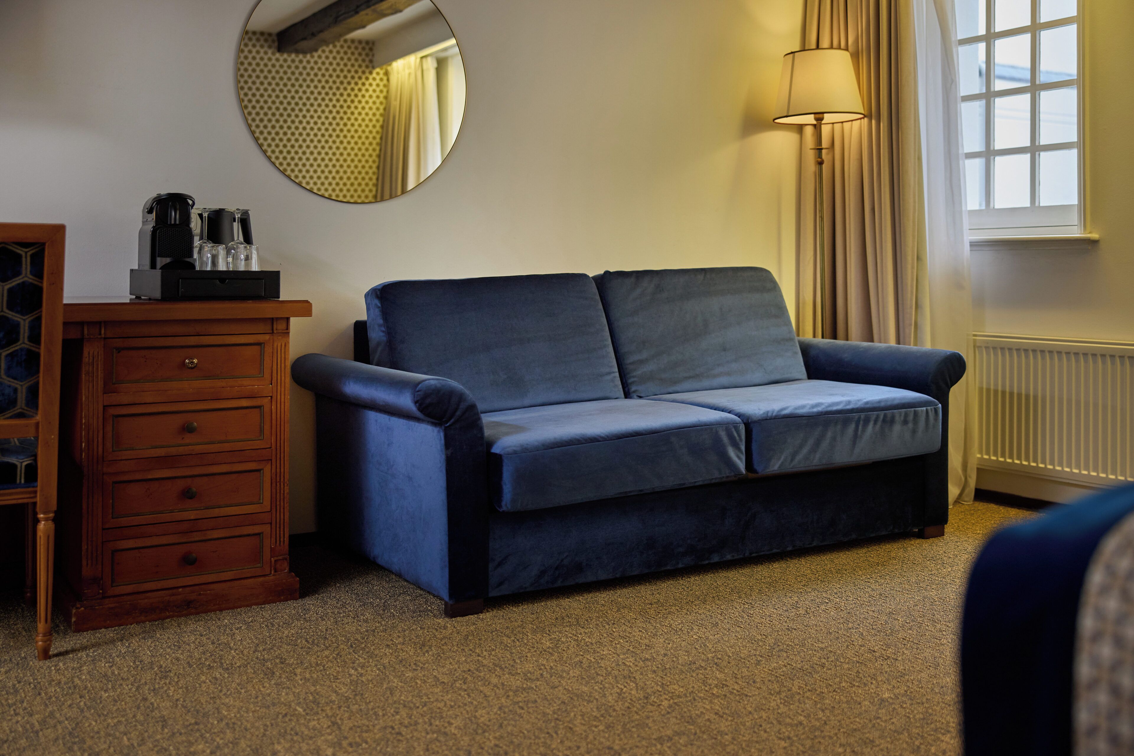 A room with a blue sofa, a wooden dresser, a mirror, a lamp, and a window with curtains.